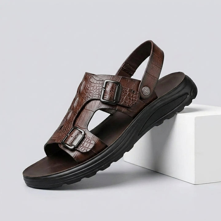 A single brown leather sandal with crocodile-embossed straps and dual buckle closures, resting on a white geometric block against a light gray background.
