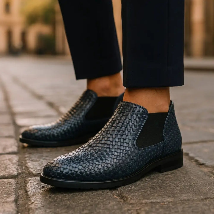 A close-up of a person wearing dark woven ankle boots with black elastic side panels, paired with dark trousers, standing on a stone pavement.