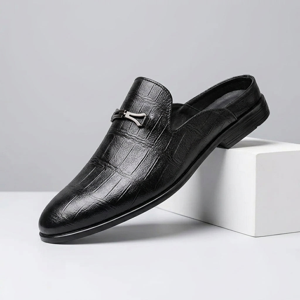 A single black crocodile-embossed leather loafer slip-on shoe with a silver horsebit detail, resting on a white geometric block against a light gray background.