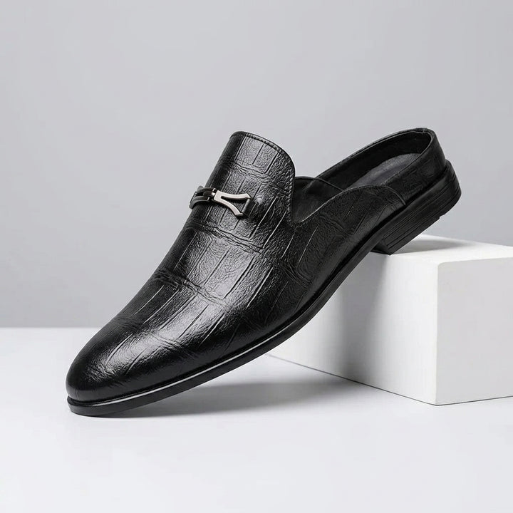 A single black crocodile-embossed leather loafer slip-on shoe with a silver horsebit detail, resting on a white geometric block against a light gray background.