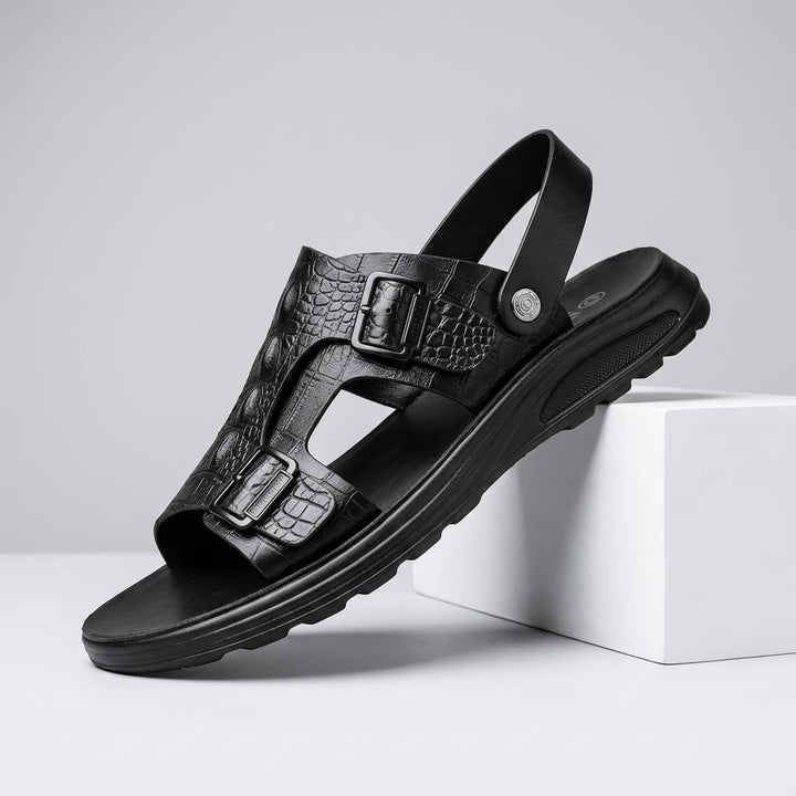 A single black leather sandal with textured crocodile-embossed straps and dual buckle closures, shown resting on a white display block against a light gray studio background.