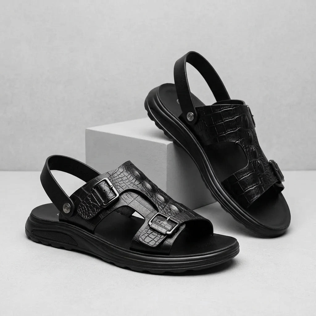 A pair of black leather sandals with crocodile-embossed straps and adjustable buckles, displayed on a light gray surface with a white block prop.