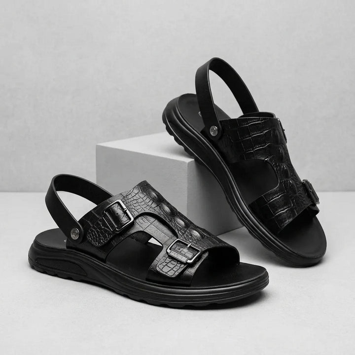 A pair of black leather sandals with crocodile-embossed straps and adjustable buckles, displayed on a light gray surface with a white block prop.