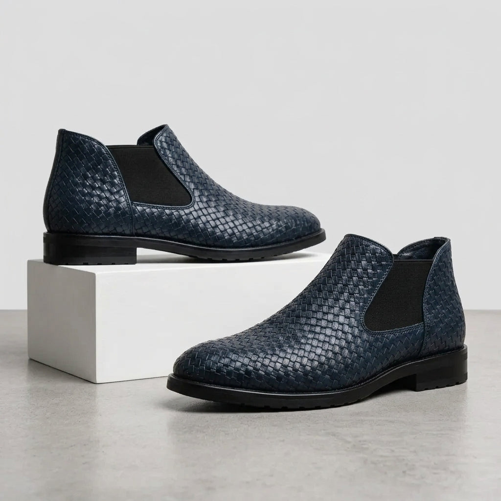 A pair of men's Chelsea boots in navy blue woven leather with black elastic side panels, displayed on a light gray floor with one boot resting on a white block.