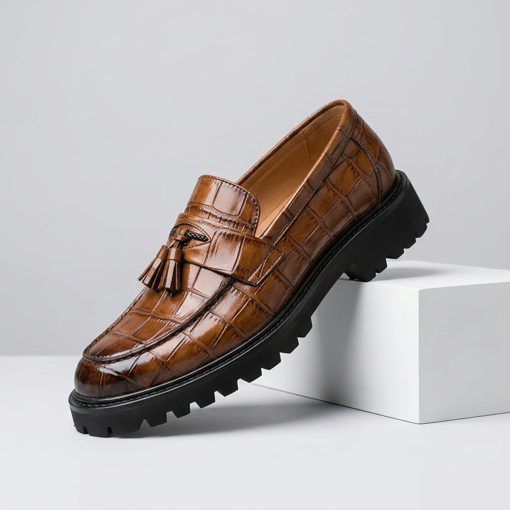 A single brown crocodile-embossed loafer with a tassel detail and a chunky black lug sole, shown resting on a white display block against a light gray background.