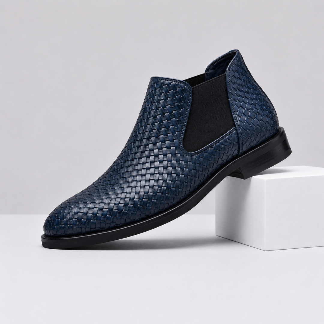A single men's ankle boot in navy blue woven leather with black elastic side panels and a black sole, displayed on a white block against a light gray studio background.