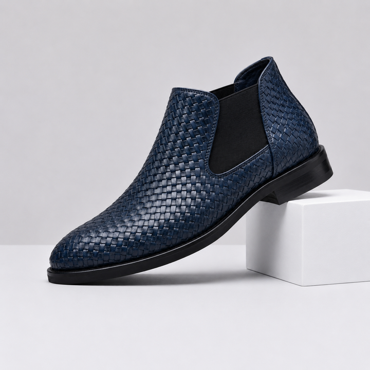 A single men's ankle boot in navy blue woven leather with black elastic side panels and a black sole, displayed on a white block against a light gray studio background.